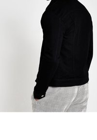 Man som bär en svart jacka med sherpa-krage och grå rutiga byxor, stående med vänster hand i fickan mot en vit bakgrund.