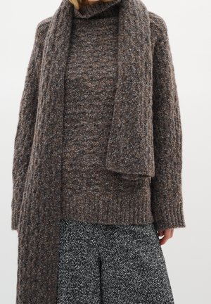 Femme portant un pull en tricot marron texturé avec une longue écharpe assortie et une jupe noire et blanche à motif chevrons.
