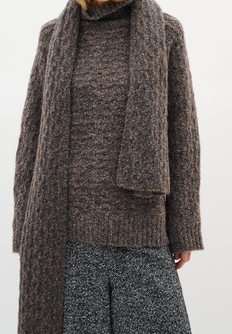 Femme portant un pull en tricot marron texturé avec une longue écharpe assortie et une jupe noire et blanche à motif chevrons.