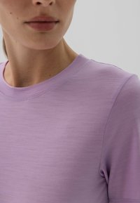 Zbliżenie osoby w gładkiej, jasnofioletowej koszulce z okrągłym dekoltem, pokazującej częściowo twarz, szyję i górną część torsu na neutralnym tle.