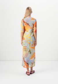 Vrouw met lang blond haar, gekleed in een kleurrijke midi-jurk met tropische print en rode platte sandalen, staande tegen een effen witte achtergrond.