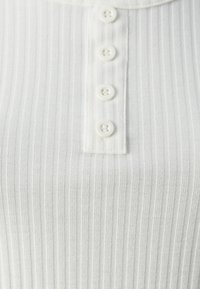 Top bianco a costine con bottoni, caratterizzato da strisce verticali, colletto piccolo e sei bottoni coordinati sul davanti.