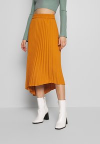 Jupe midi à plis en jaune moutarde avec une taille élastique, associée à des bottines blanches en cuir crocodile à talon bloc.