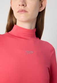 Roza majica s podignutim ovratnikom napravljena od glatkog materijala, s srebrnim logotipom "tommy jeans" na sredini. Prilegajući dizajn i bešavna tekstura.