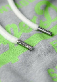 Felpa grigia con grande testo neon verde crepato e cappuccio con stringhe bianche. Le estremità delle stringhe sono in metallo argentato con dettagli di branding incisi.