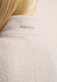 Hellbeige strukturierte Jacke mit hohem Kragen und silbernem "bugatti"-Logo am hinteren Halsausschnitt. Weicher Stoff mit einem gewebten Muster.