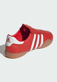 Crvene kožne sportske cipele s gumenenim potplatom, bijele tri pruge i čičak zatvaranjem. Glatka tekstura i zaobljen dizajn vrha.
