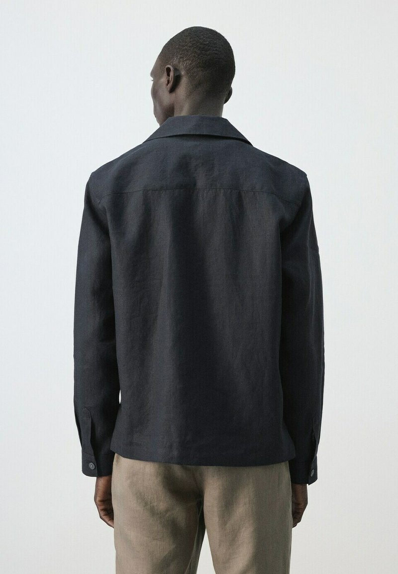 Homme vu de dos portant une chemise noire à manches longues et un pantalon beige sur un fond uni de couleur claire.