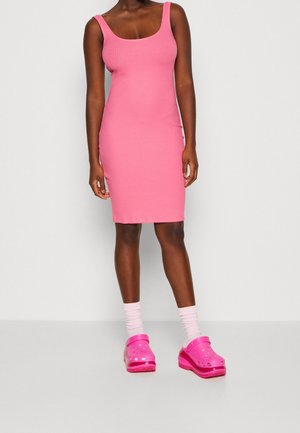 Vrouw draagt een mouwloze geribbelde roze jurk tot op de knie, met lichtroze sokken en felroze klompen tegen een effen achtergrond.