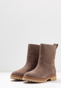 Bottines en daim marron à texture lisse, bout arrondi et semelle extérieure en caoutchouc. Comprend des accents cousus et un logo sur le côté.