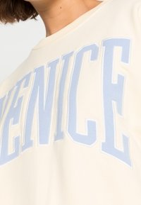 Felpa crema con grande scritta "VENICE" di colore azzurro chiaro sul davanti, realizzata in tessuto morbido, con scollatura rotonda e vestibilità casual.