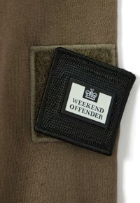 Patch "Weekend Offender" in bianco e nero con logo a corona attaccato a tessuto marrone mediante un patch in velcro quadrato di colore verde oliva.