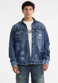 Denim jack met een lichtblauwe wassing, versleten details, twee borstzakken en een knoopsluiting aan de voorzijde. Daaronder is een wit T-shirt met een grafische print.
