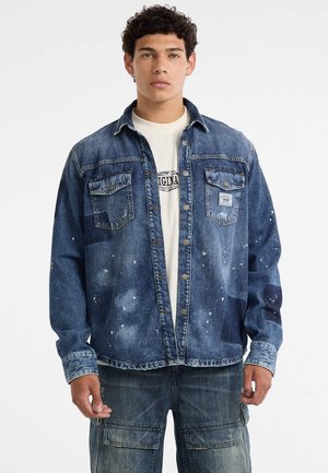 Denim jack met een lichtblauwe wassing, versleten details, twee borstzakken en een knoopsluiting aan de voorzijde. Daaronder is een wit T-shirt met een grafische print.
