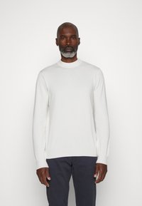 Pull en tricot blanc à manches longues avec un col rond, fabriqué à partir d'un tissu doux, associé à un pantalon sombre. Design minimaliste, texture lisse.