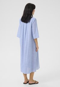 Abito azzurro chiaro, lungo fino al ginocchio, realizzato in tessuto morbido, con maniche a tre quarti, una piega sulla schiena e una vestibilità rilassata. Indossato con infradito nere.