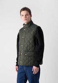 Gilet imbottito verde con colletto alto, bottoni a pressione dorati e due tasche frontali, indossato sopra una camicia nera a maniche lunghe e jeans blu.