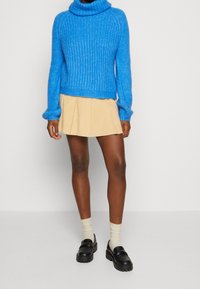 Personne portant un pull à col roulé bleu vif, une mini-jupe plissée beige, des chaussettes crème et des mocassins noirs épais sur un fond blanc.