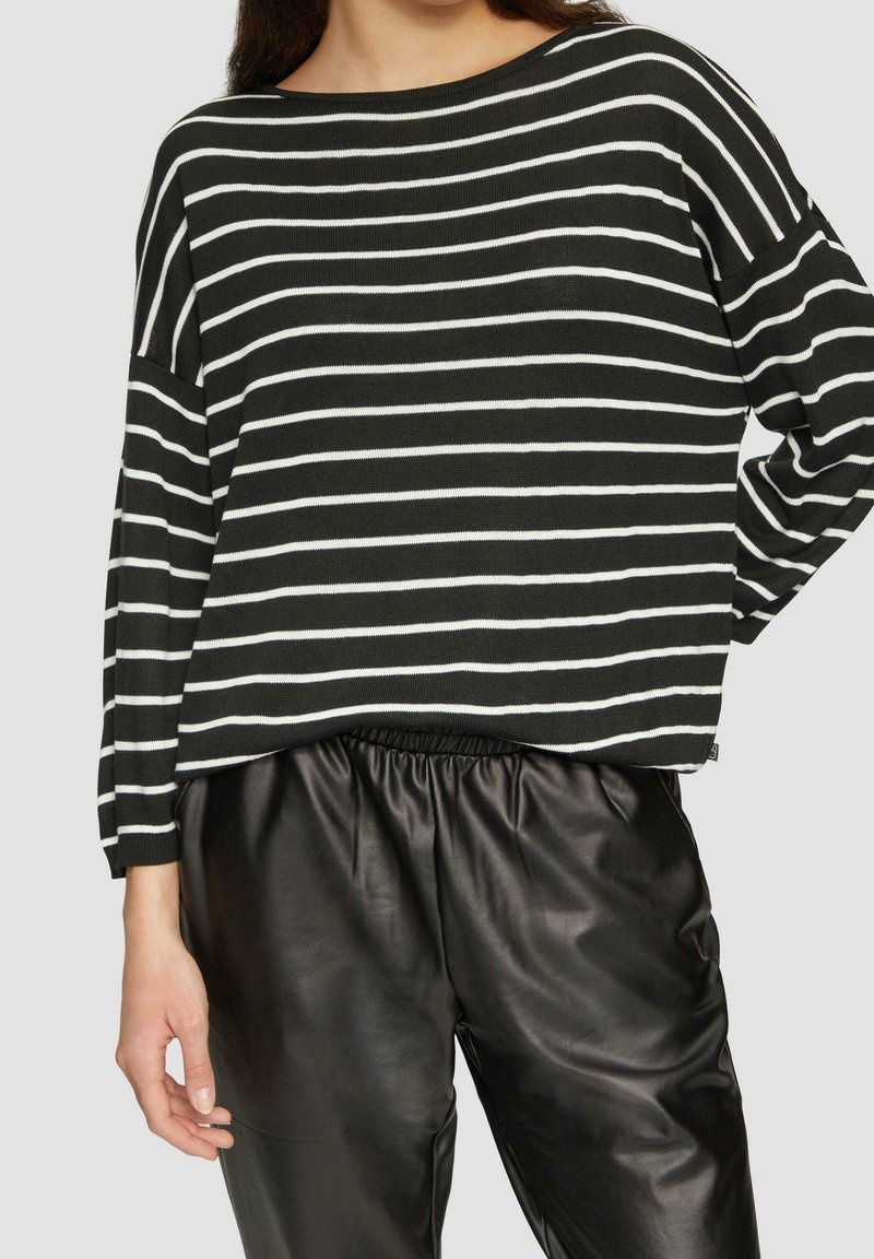 Femme portant un haut à manches longues rayé noir et blanc horizontalement et un pantalon noir en simili-cuir avec ceinture élastique, debout devant un fond uni.