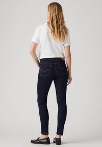 Jeans ajustados de color azul oscuro con costuras amarillas, que presentan un diseño clásico de cinco bolsillos y un emblema en el bolsillo trasero. Llevados con una camiseta blanca y zapatos negros.