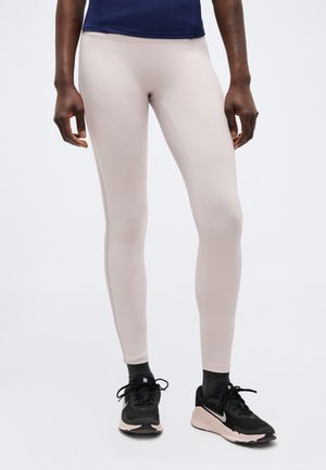 Osoba w beżowych legginsach, czarnych butach Nike z różowymi podeszwami, ciemnych skarpetkach i granatowym topie, stojąca na białym tle.