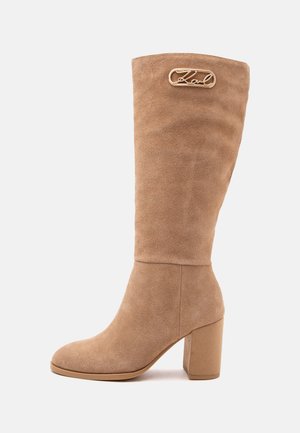 Botte en daim beige haute avec talon bloc et détail du logo en métal doré près du haut, présentée en profil latéral sur fond blanc.