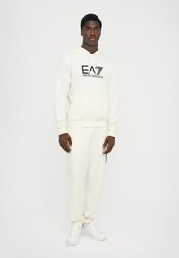 Set di felpa color crema e pantaloni joggers, in materiale felpato, con logo EA7 nero. Il modello è in piedi su uno sfondo bianco, indossando sneaker bianche.