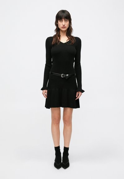 Schwarzes, geripptes Strickkleid mit langen Ärmeln, V-Ausschnitt und ausgestelltem Rock; kombiniert mit einem schwarzen Gürtel, schwarzen kniehohen Socken und High Heels.