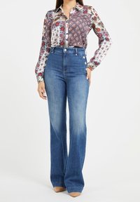 Kvinna i högmidjade, utsvängda blå jeans med sidoknappar och en långärmad blommig skjorta i lappteknik, stående mot vit bakgrund.