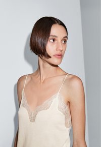 Beige hemdje met kantafwerking, voorzien van dunne bandjes en een zachte textuur. Het kant heeft een bloemenpatroon en een golvende rand.