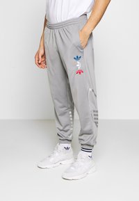 Muž oblečený v šedých adidas teplácích se modrými, bílými a červenými logy trefoil, v bílých teniskách a v ponožkách adidas, stojící na bílém pozadí.