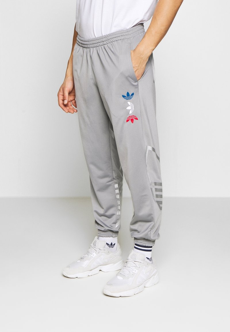 Muž oblečený v šedých adidas teplácích se modrými, bílými a červenými logy trefoil, v bílých teniskách a v ponožkách adidas, stojící na bílém pozadí.