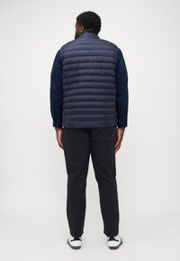 Gilet matelassé bleu marine avec un design quilté, col montant et matériau léger, associé à un pantalon sombre et des chaussures de sport blanches.