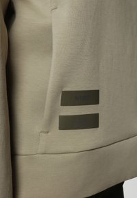 Sudadera beige con una textura suave, que presenta dos etiquetas de metal oscuro que muestran "N1987" y "PROPIEDAD DEGRADADA ACEPTABLE."