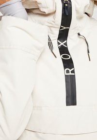 Cremefarbener Windbreaker mit einem auffälligen schwarzen Roxy-Logo auf der Vorderseite, ausgestattet mit einem Reißverschlusskragen, Kordelzügen und gemusterten Akzenten an den Schnüren.