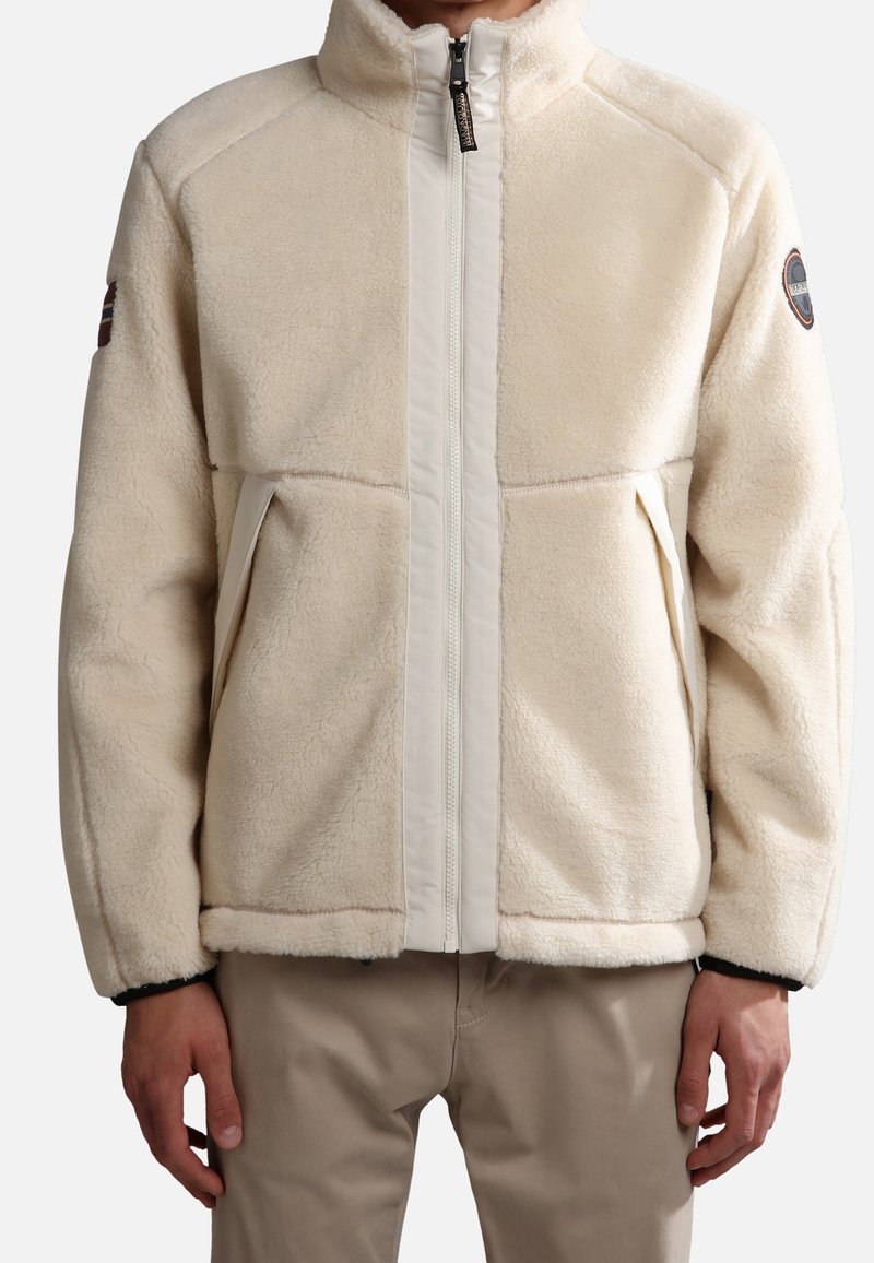Beige fleece jacket with a high collar, full front zip, and side pockets. Features contrast fabric panels and a round logo patch on the sleeve.