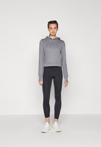 Graue, kurze Kapuzenjacke mit verstellbarem Kapuzenzug, kombiniert mit schwarzen Leggings und weißen Turnschuhen. Der Stoff hat eine glatte Textur ohne Muster.