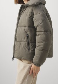 Person in olivgrüner gepolsterter Jacke mit Frontreißverschluss und seitlicher Reißverschlusstasche, kombiniert mit beigen Cordhosen.