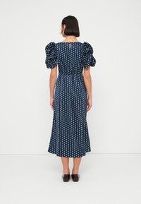 Robe bleu marine à pois avec manches bouffantes, corsage ajusté et jupe longueur cheville. Texture lisse avec une ouverture en forme de goutte d'eau dans le dos.