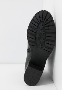 Semelle de botte noire avec des crampons profonds et des motifs en forme de croix, placée à la verticale contre un mur d'angle blanc et un sol blanc.