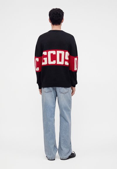 Personne portant un pull noir et rouge avec les lettres blanches GCDS sur le dos et un jean large bleu clair, debout de dos.