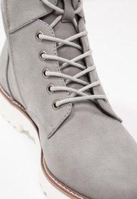 Bottine en suède gris avec six œillets en métal et lacets gris. Dispose d'une semelle en caoutchouc blanche à bande de roulement texturée et d'un accent marron contrastant.