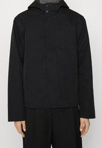 Black waterproof jacket with a hood, featuring a front snap closure, a chest pocket, and long sleeves. Smooth texture and minimalist design.