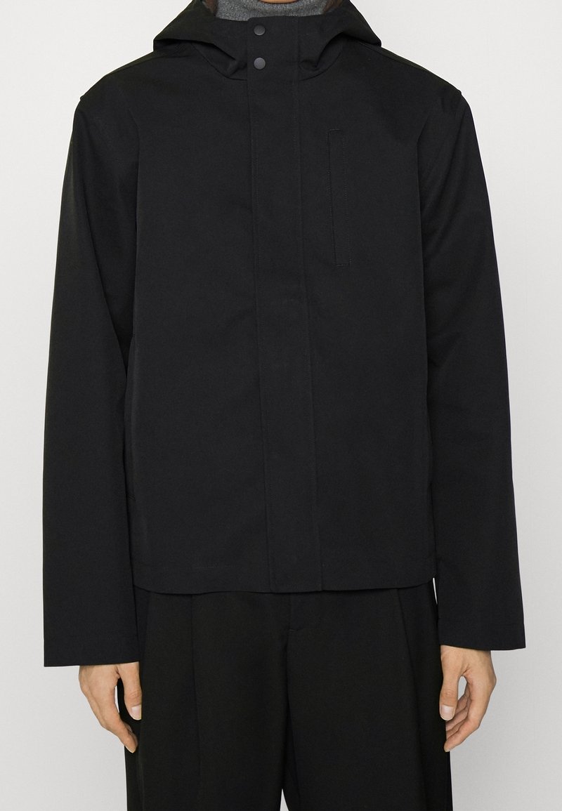 Black waterproof jacket with a hood, featuring a front snap closure, a chest pocket, and long sleeves. Smooth texture and minimalist design.