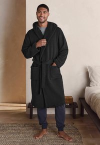 Rocă neagră cu glugă, având o căptușeală texturată la interior, legată pe față și două buzunare frontale. Asortată cu pantaloni de trening bleumarin și picioare goale.