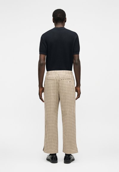 Homme portant un pull en maille à manches courtes noir, pantalon large à carreaux beige avec poches arrière, et chaussures habillées noires, tourné de dos sur fond blanc.