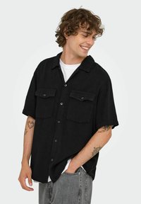 Sort kortærmet skjorte med knaplukning, to frontlommer og en afslappet pasform. Båret over en hvid t-shirt med synlige grå jeans.