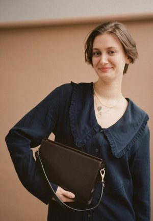 Camicia blu navy con colletto arricciato; tiene una pochette in pelle marrone scuro con strap e chiusura; molte collane a strati.
