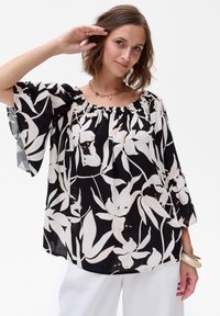 Czarna bluzka w białe wzory kwiatowe, z marszczonym dekoltem i dzwonowymi rękawami. Lekki materiał o luźnym kroju, zestawiona z białymi spodniami.