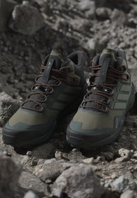 Par de zapatos de senderismo verde oscuro y negro con cordones naranjas descansando sobre terreno rocoso.